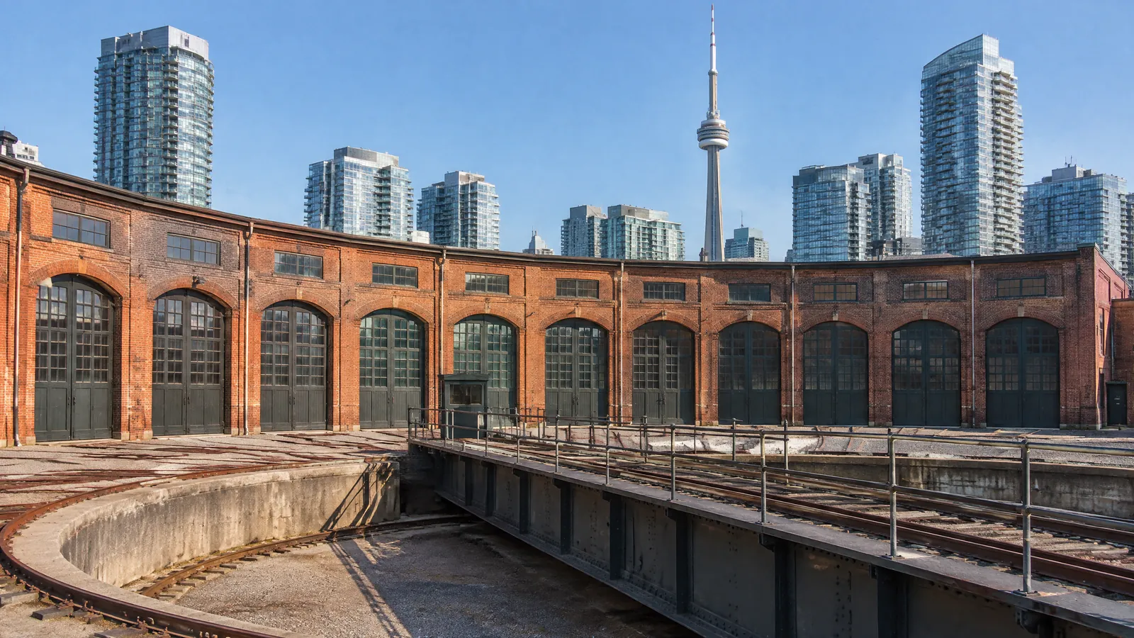 Curved brick railway roundhouse with arched doors and a locomotive turntable in downtown Toronto on a sunny day, the CN Tower and condo towers behind